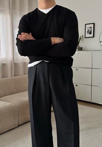 Pull noir à col en V superposé sur une chemise blanche, associé à un pantalon noir plissé taille haute. Tissu souple, coupe décontractée.