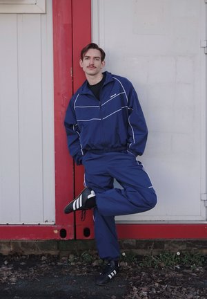 Hombre con bigote, vestido con chándal azul y zapatillas negras Adidas, apoyado contra una pared roja y blanca, de pie sobre una pierna con las manos en los bolsillos.