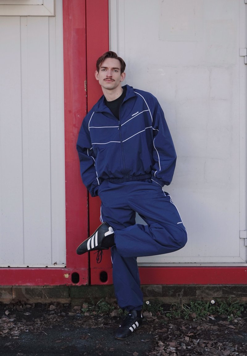 Hombre con bigote, vestido con chándal azul y zapatillas negras Adidas, apoyado contra una pared roja y blanca, de pie sobre una pierna con las manos en los bolsillos.