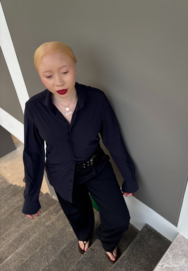 Persoon met lichte huid en blond haar, gekleed in zwart en met rode lippenstift, staande op tapijtbeklede trap tegen een grijze muur.