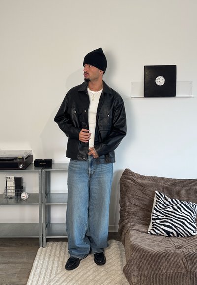 Chaqueta de cuero negra con dos bolsillos delanteros, camiseta blanca, jeans azules holgados y botas negras. Gorro de punto negro. Cojín con patrón de cebra.