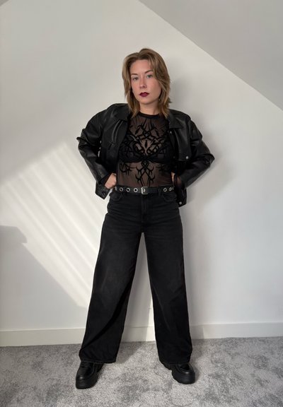 Chaqueta de cuero negra sobre una blusa negra transparente con patrones intrincados, combinada con jeans de pierna ancha de talle alto y zapatos negros gruesos.