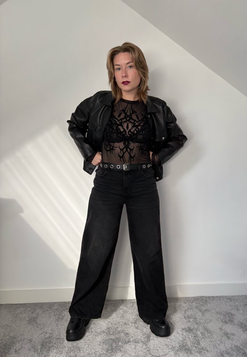 Black leather jacket over a sheer black top with intricate patterns, paired with high-waisted black wide-leg jeans and chunky black shoes.