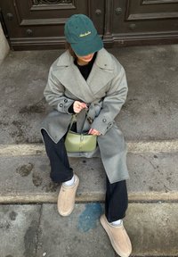 Grahamfarbener Wollmantel mit großem Kragen, schwarzer Strickpullover, schwarze Jeans, beige UGG Hausschuhe und eine grüne Kappe. Hält eine kleine grüne YSL Handtasche.