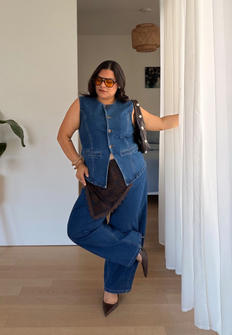Femme portant un gilet en jean sans manches, un pantalon en jean, des talons marron et des lunettes de soleil orange, debout à l'intérieur près de rideaux blancs avec une jambe croisée.