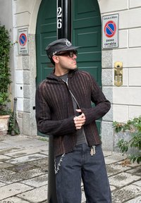 Cardigan marrone a coste con cappuccio, apertura anteriore e polsini sfrangiati. Abbinato a jeans scuri con decorazioni in catena argentata.