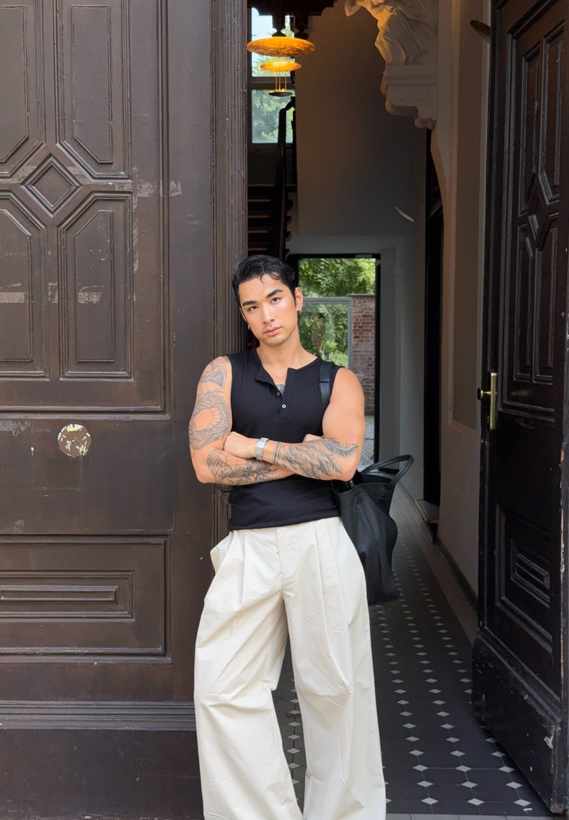 Junge tätowierte Person in schwarzem ärmellosem Shirt und weißen weiten Hosen steht mit verschränkten Armen im Eingang eines kunstvollen Gebäudes.