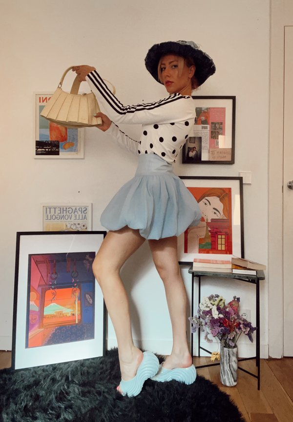 Woman wearing a black hat, polka dot top, blue puff skirt, and sculpted shoes poses holding a beige handbag in a room with framed art and flowers.