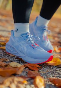 Zapatillas de running azul claro con parte superior de malla texturizada, detalles rojos en la suela y cordones planos, colocadas sobre una superficie de hojas de otoño y grava.