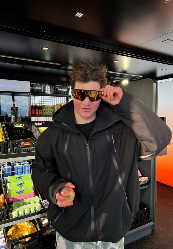 Jeune personne en veste noire ajustant des lunettes de soleil réfléchissantes, tenant une pomme, debout dans un magasin avec des étagères de produits alimentaires en arrière-plan.