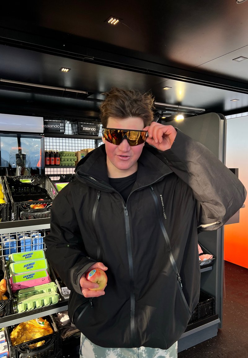 Giovane con giacca nera che si sistema occhiali da sole riflettenti, tiene in mano una mela, in piedi in un negozio con scaffali di alimenti sullo sfondo.