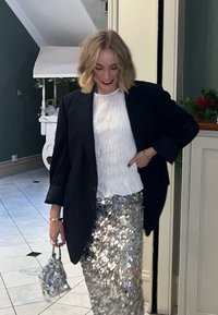 Blazer noir surdimensionné, haut blanc texturé et jupe argentée à sequins. Petit sac à main argenté. Arrière-plan avec des murs vert foncé et un sol en marbre.