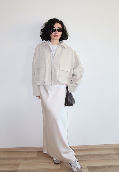 Mujer con cabello rizado que lleva gafas de sol oscuras, chaqueta beige oversize, falda larga color crema, zapatillas deportivas y sostiene un pequeño bolso oscuro.