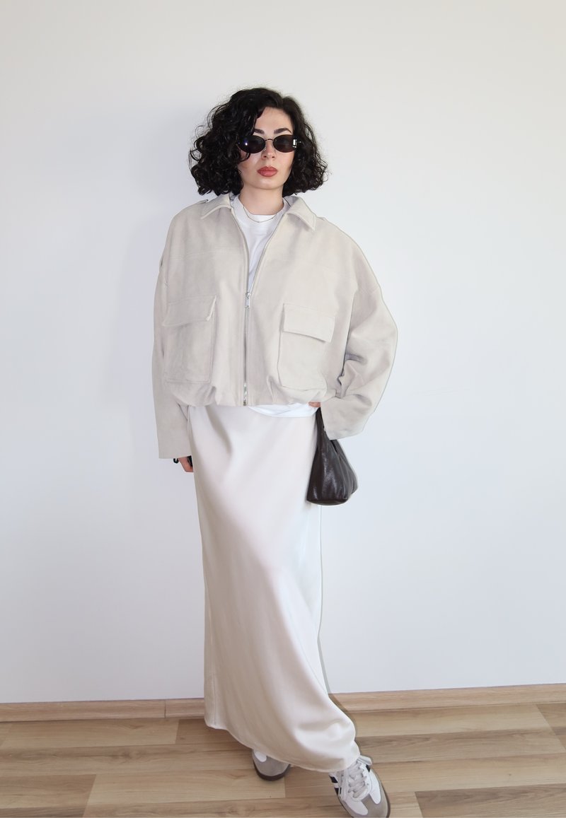 Göndör hajú nő sötét napszemüvegben, nagy méretű bézs dzsekiben, hosszú krémszínű szoknyában, tornacipőben, és egy kis sötét kézitáskát tartva.