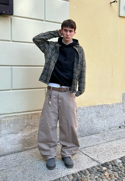 Pantalones beige de gran tamaño con un acabado texturizado, combinados con una sudadera negra y una camisa a cuadros verdes. Zapatos grises y un cinturón con llavero completan el conjunto.