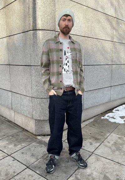 Camisa de franela a cuadros en verde y rosa, camiseta gráfica gris con diseño de perro, jeans oscuros, gorro gris, zapatillas gris y negro.