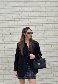 Γυναίκα με μακριά μαλλιά που φοράει μαύρα γυαλιά ηλίου, μαύρο σακάκι, καρό φούστα και κρατάει μαύρη τσάντα, στέκεται μπροστά σε έναν ανοιχτόχρωμο τοίχο από τούβλα.