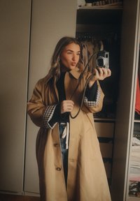 Lang beige trenchcoat med vide ermer, med en sort genser under og rutete mansjetter. Frakken har en avslappet passform og knapppdetaljer.