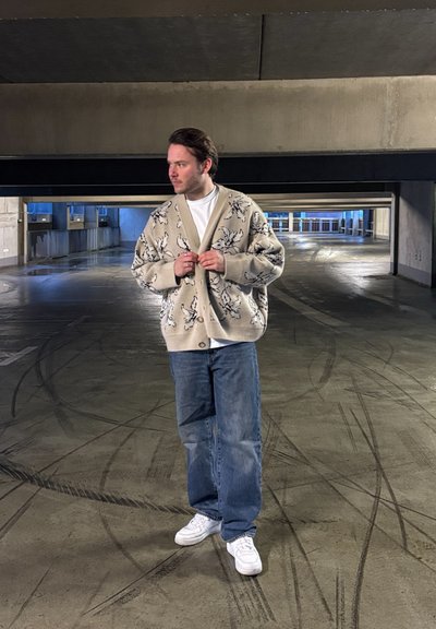Cárdigan beige con patrones florales, de corte oversize, con botones en la parte delantera. Llevado sobre una camiseta blanca y combinado con jeans azules holgados y zapatillas blancas.
