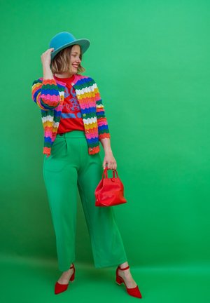 Mujer con cárdigan de rayas multicolores, camisa roja, pantalones verdes, sombrero azul, zapatos rojos, sosteniendo un bolso rojo, de pie frente a un fondo verde, sonriendo.
