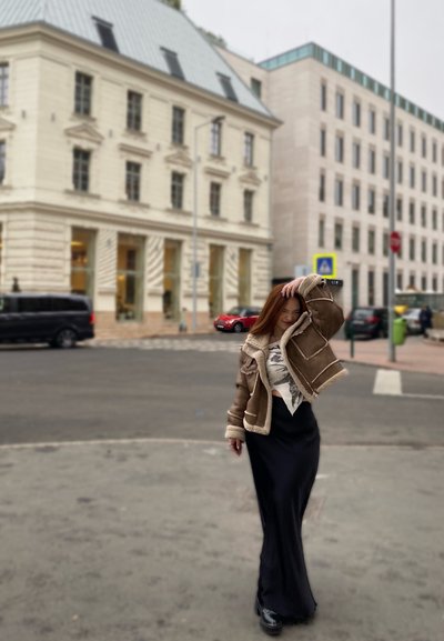 Chaqueta de borrego marrón con costuras blancas sobre una blusa estampada, combinada con una falda larga negra y botas negras gruesas. Entorno urbano.