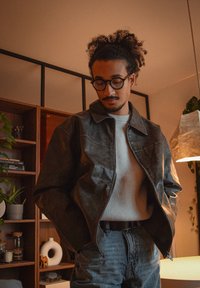 Jeune homme aux cheveux bouclés et lunettes portant une veste en cuir et un pull, debout à l'intérieur près d'une étagère en bois et d'une lampe.
