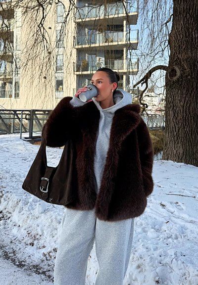 Mujer con sudadera gris y abrigo de piel bebe de un vaso de papel al aire libre en una zona urbana nevada, llevando un bolso grande y oscuro al hombro.