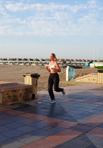 Mujer corriendo por un camino pavimentado con un sujetador deportivo blanco y mallas negras, usando zapatillas de running azul claro. En el fondo hay sombrillas de playa.