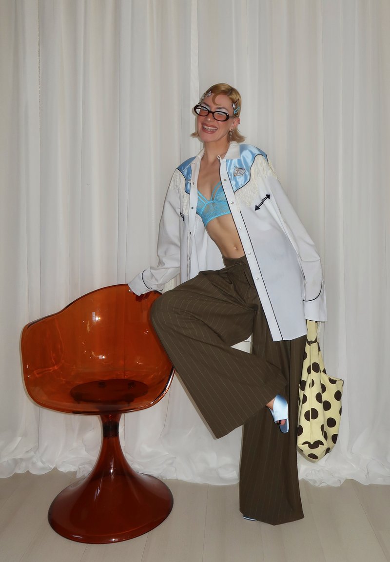 Personne souriante, portant des lunettes, un bralette bleu, une veste blanche oversize, un pantalon large marron, tenant un sac à pois, appuyée sur une chaise orange.