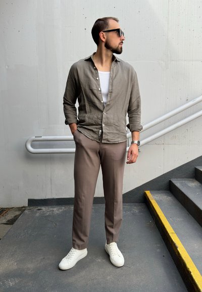 Camisa de lino gris claro, camiseta blanca de tirantes, pantalones marrones y zapatillas blancas. Los accesorios incluyen gafas de sol y un reloj. Entorno urbano con escaleras.