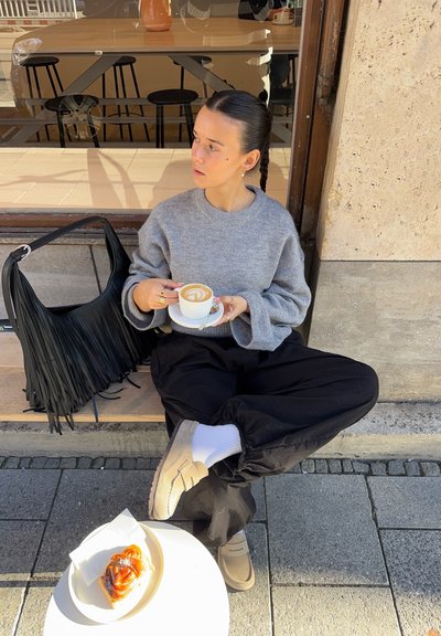 Suéter gris, pantalones holgados negros, zapatos tipo slip-on beige con calcetines blancos. Sosteniendo una taza de café con arte latte. Bolso negro con flecos cerca. Pastel en un plato.