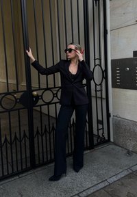 Femme en costume noir et lunettes de soleil appuyée contre une grille en fer forgé noire près d'une entrée de bâtiment en pierre avec un panneau d'interphone.