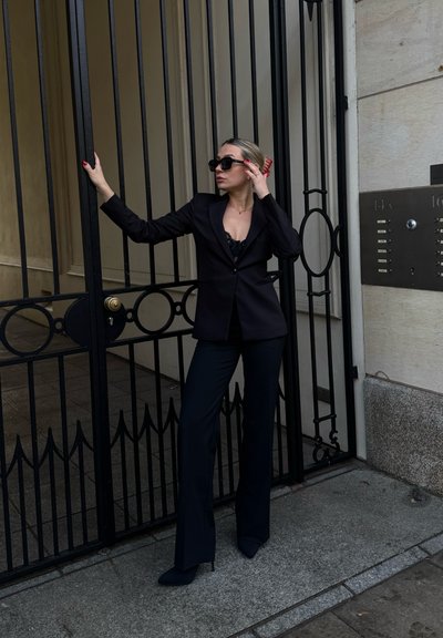 Mujer con traje negro y gafas de sol apoyada en una puerta de hierro forjado negra cerca de la entrada de un edificio de piedra con un panel de intercomunicación.