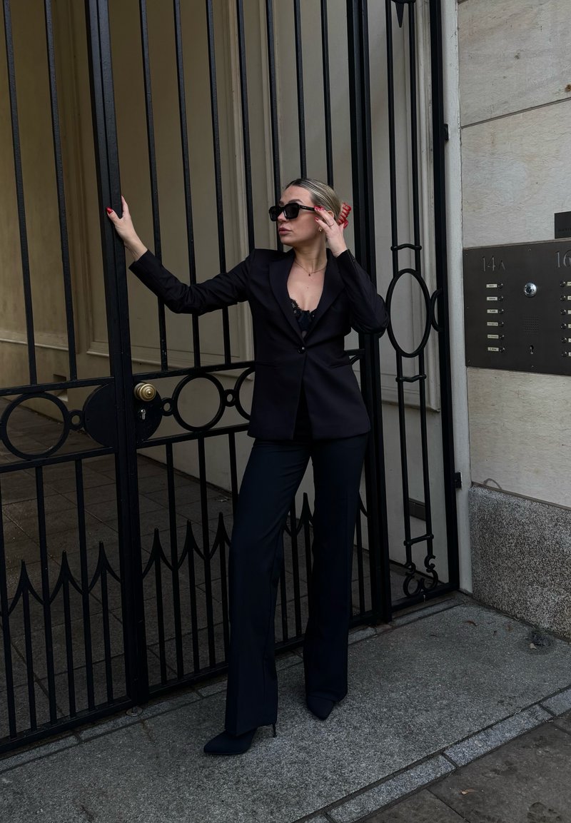 Femme en costume noir et lunettes de soleil appuyée contre une grille en fer forgé noir près de l'entrée d'un bâtiment en pierre avec un panneau d'interphone.
