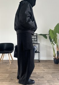 Čierna nadýchaná bomber bunda s rebrovanými manžetami, nosená na čiernych širokých nohaviciach s bočným vreckom. Matný povrch, pohodlný strih.