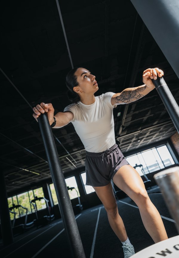 Frau mit tätowiertem Arm, die ein weißes Shirt und schwarze Shorts trägt, trainiert drinnen und hält zwei vertikale Stangen im Fitnessstudio mit großen Fenstern.