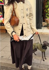 Personne en veste beige avec des épingles, jupe noire et sac à main marron tenant la laisse d'un petit chien en manteau vert sur un trottoir en ville.