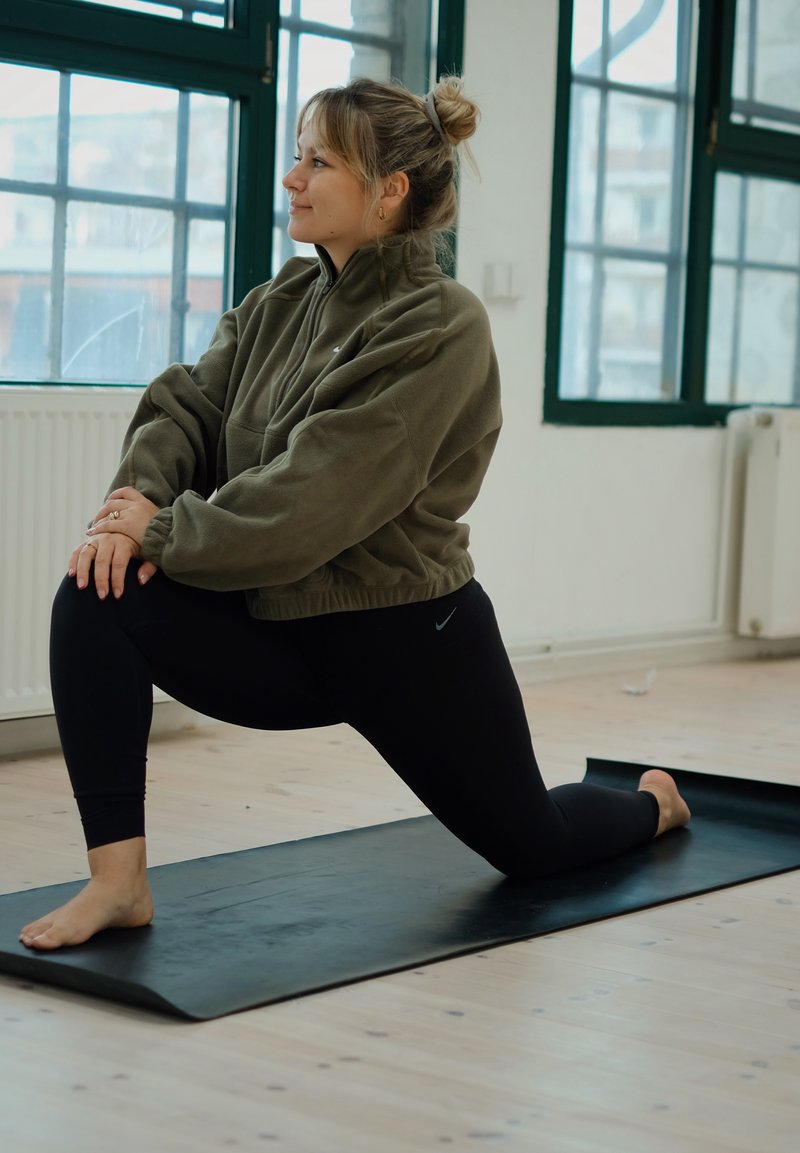 Tapis de yoga noir sur un sol en bois, avec une personne en veste polaire verte et leggings noirs effectuant un étirement en fente. Lumière naturelle provenant des fenêtres.