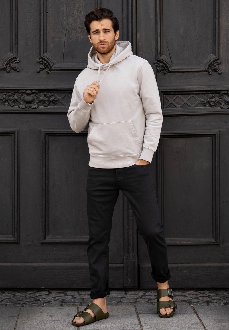 Lichtgrijze hoodie met buidelzak, zwarte slim-fit jeans en donkergroene sandalen met verstelbare banden, tegen een donkere houten achtergrond.