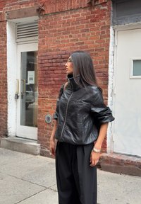 Chaqueta recortada de cuero negro con un alto cuello y cremallera frontal, combinada con pantalones negros holgados. El fondo presenta una pared de ladrillo y puertas.