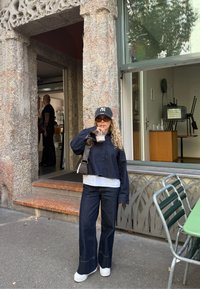 Kvinna som bär mörka jeans, marinblå jacka och keps dricker iskaffe utanför stenbyggnad nära grönt bord och metallstolar.