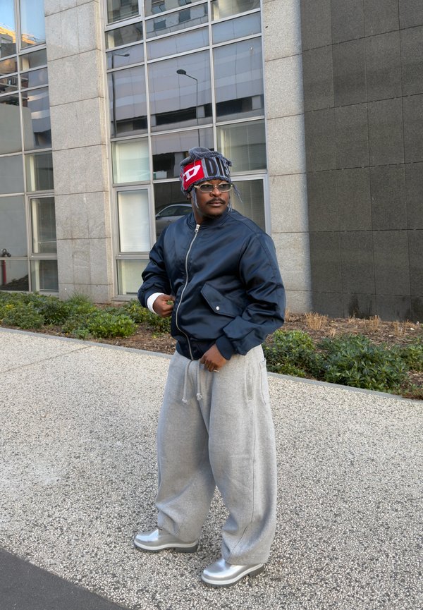 Man wearing a navy bomber jacket, grey wide sweatpants, silver shoes, glasses, and a colourful beanie stands on a city pavement near a building.