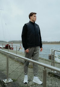 Črna izolirana usnjena jopica čez črno majico z dolgimi rokavi, v kombinaciji s svetlo sivimi trenirka hlačami in belimi športnimi copati. Zunanji prostor ob vodi.