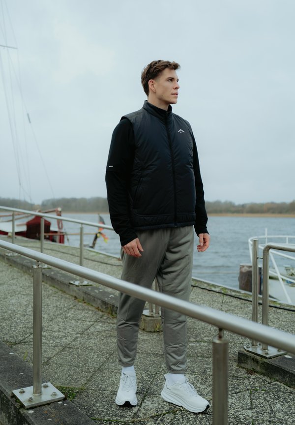 Chaleco aislante negro sobre una camiseta de manga larga negra, combinado con pantalones de chándal gris claro y zapatillas blancas. Entorno al aire libre junto al agua.