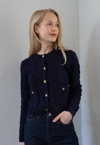 Tmavomodrý pletený kardigán so vzormi vrkočov, rebrovanými manžetami a dvoma prednými vreckami. Má zlaté gombíky a okrúhly výstrih.