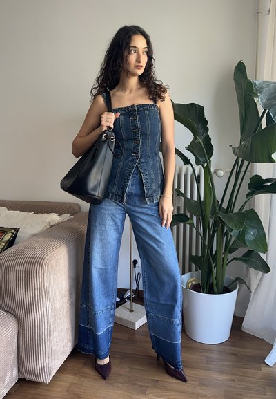 Mujer con cabello rizado que lleva un top vaquero sin tirantes y jeans de pierna ancha, sosteniendo un bolso negro y de pie junto a una gran planta de interior.