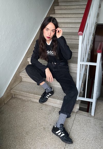 Sudadera negra de Adidas con logo, jeans negros holgados, calcetines grises y zapatillas negras con rayas blancas, sentado en escalones de piedra cerca de una barandilla.