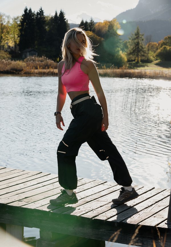 Vrouw in een roze crop top en zwarte broek die op een houten steiger staat naast een meer met een bos en bergen op de achtergrond tijdens zonsondergang.
