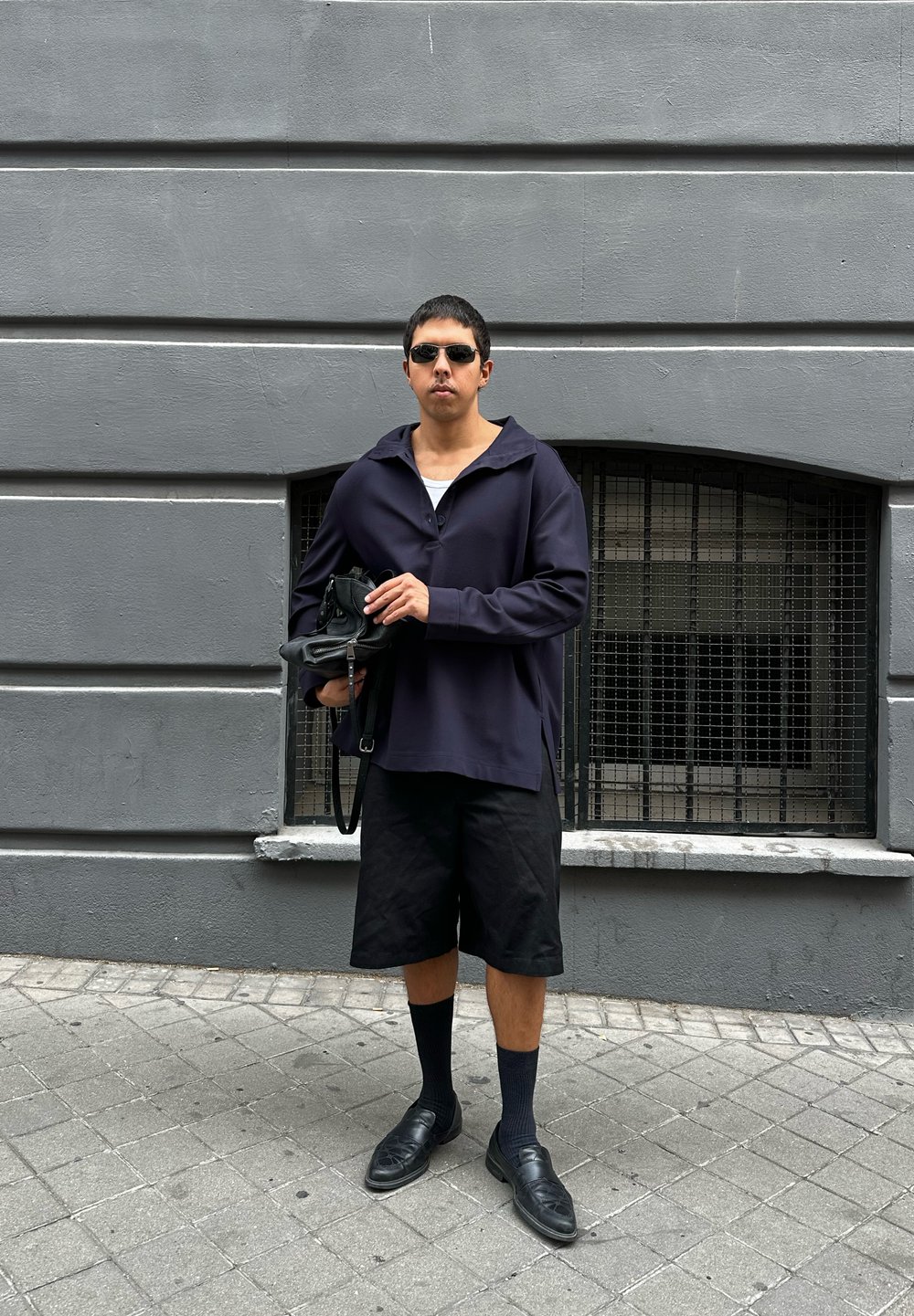 Pullover oversize blu navy con ampio colletto, pantaloni corti neri, calze nere e scarpe; tiene una piccola borsa nera contro un muro grigio.