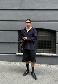 Pullover oversize blu navy con ampio colletto, pantaloni corti neri, calze nere e scarpe; tiene una piccola borsa nera contro un muro grigio.